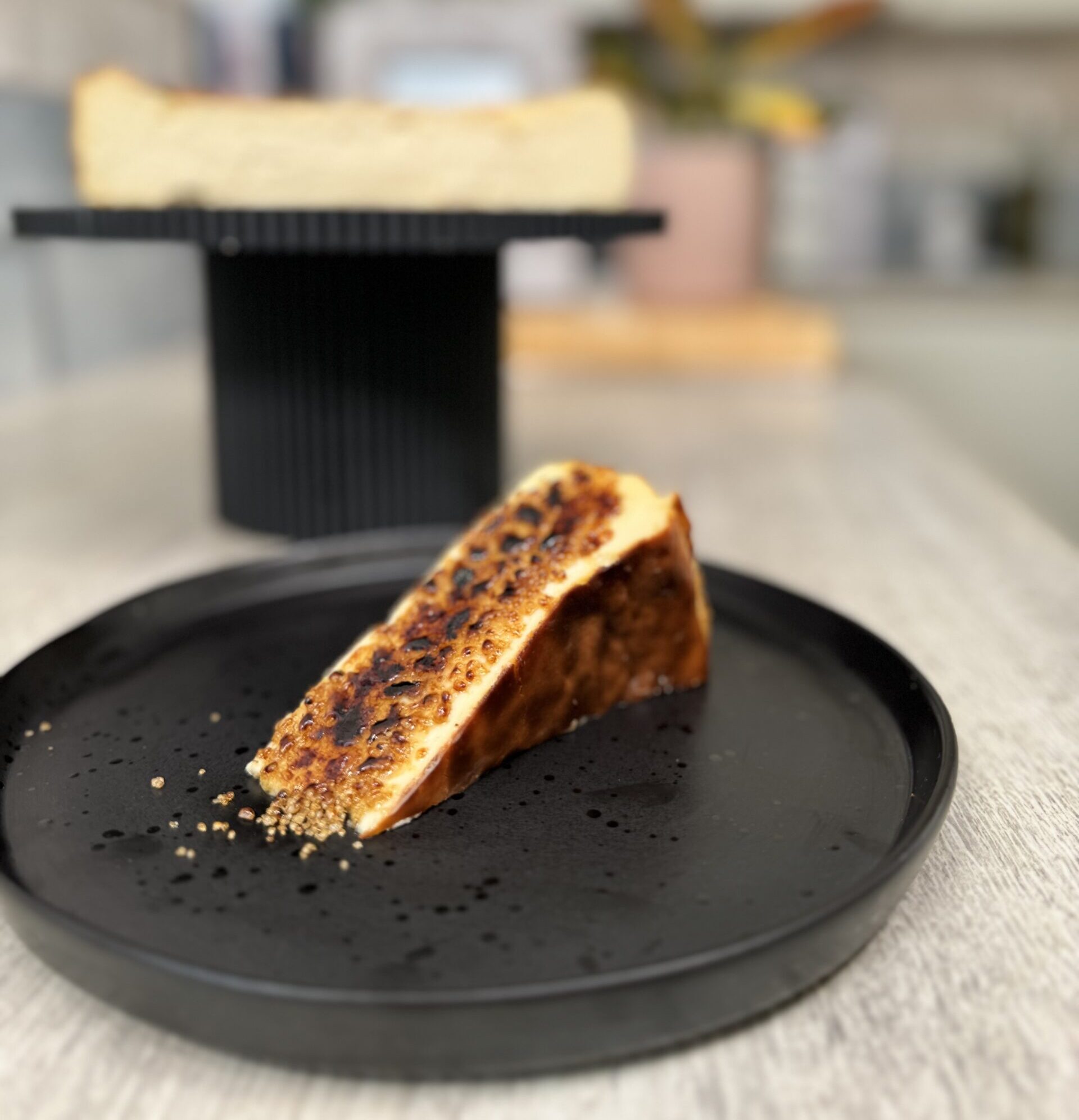 Slice of the basque cheesecake topped with caramalised sugar served on a black plate with the rest of the basque in the background