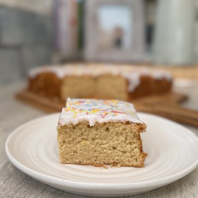 Square of vanilla cake topped with thick white icing and 100's and 1000's sprinkles with the rest of the cake behind.