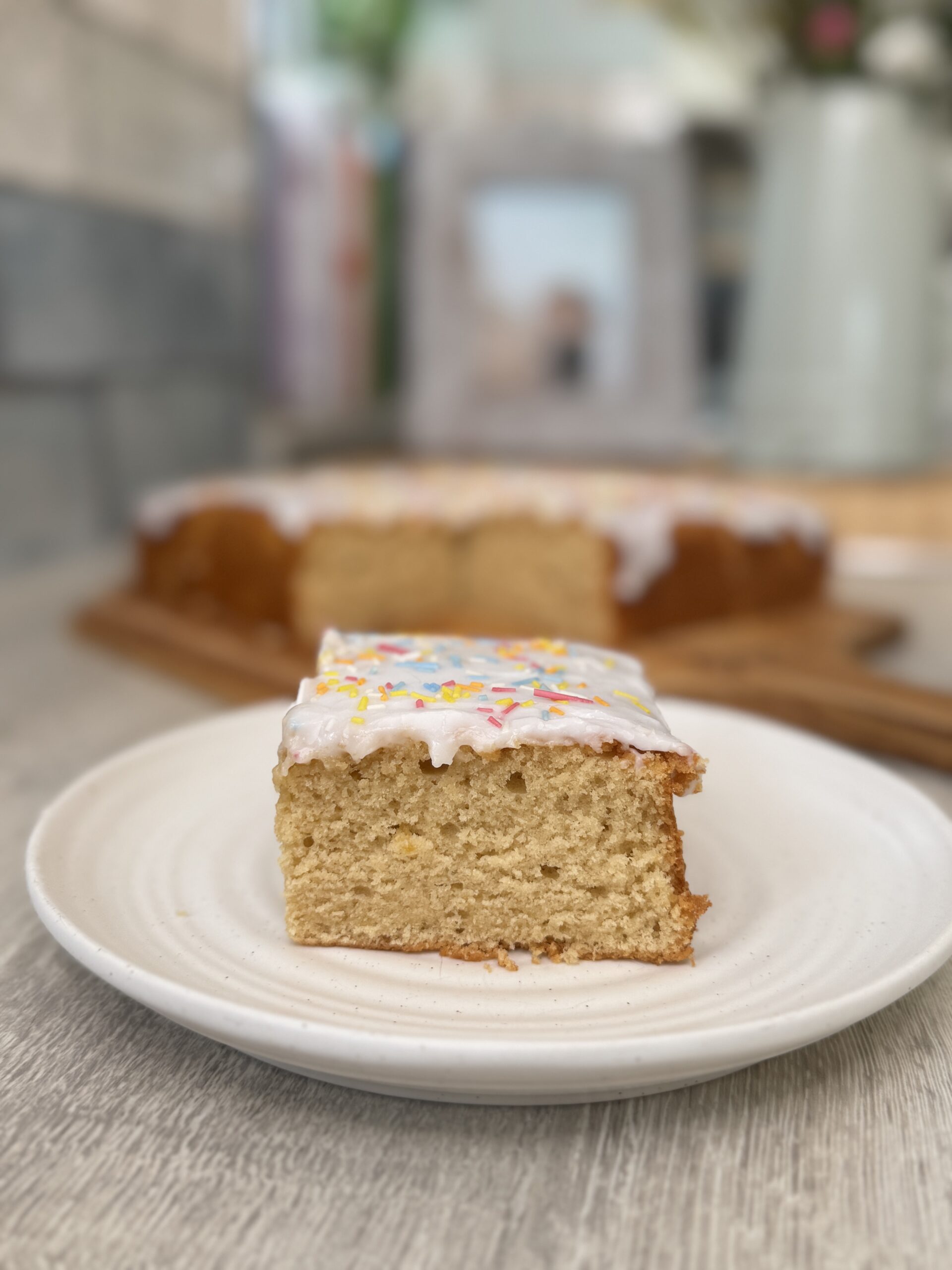 Square of vanilla cake topped with thick white icing and 100's and 1000's sprinkles with the rest of the cake behind.