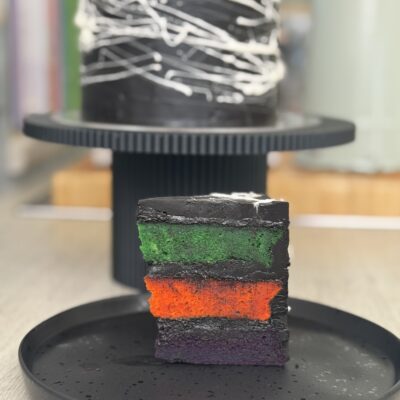 Slice of cake showing purple orange and green layers with web decoration cake behind it